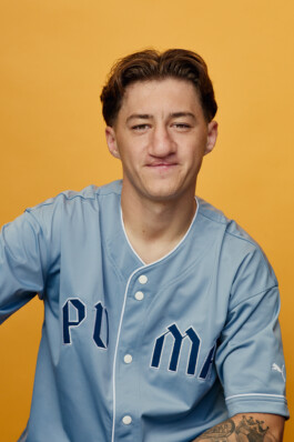 Angelo Stiller poses for a portrait at Stuttgart's MHP Arena, wearing a light blue Puma baseball-style shirt against a vibrant yellow background, his left arm with a visible tattoo resting on his knee. Photo by Roderick Aichinger for Sports Illustrated Germany.