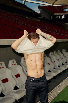 Angelo Stiller lifts his shirt over his head, revealing his toned torso and tattooed arm, while standing in front of the team benches at Stuttgart's MHP Arena. The seating area features white Recaro chairs with red accents. Photo by Roderick Aichinger for Sports Illustrated Germany.