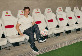 Angelo Stiller sits on the white team bench at Stuttgart's MHP Arena, dressed in a white Puma shirt and dark pants, balancing a colorful football on his foot. The background features a line of empty Recaro seats with red accents. Photo by Roderick Aichinger for Sports Illustrated Germany.