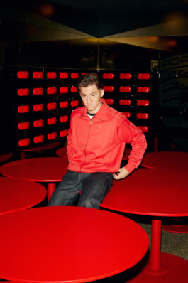 Angelo Stiller sits on a cluster of bold red circular tables, wearing a bright red Puma zip-up jacket and dark jeans. The modern interior, with a wall of red and black geometric shapes, creates a vibrant setting at Stuttgart's MHP Arena. Photo by Roderick Aichinger for Sports Illustrated Germany.
