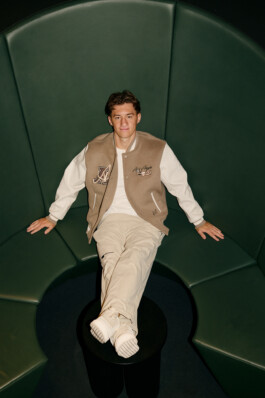 Angelo Stiller reclines on a curved green bench with his feet up on a round table, dressed in light-colored Puma clothing and a beige varsity jacket. The modern seating area and relaxed pose create a casual, contemporary atmosphere at Stuttgart's MHP Arena. Photo by Roderick Aichinger for Sports Illustrated Germany.