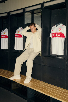Angelo Stiller poses in the team locker room at Stuttgart's MHP Arena, wearing an all-cream outfit including a Puma jacket, trousers, and sneakers. Behind him are displayed VfB Stuttgart football jerseys, creating a sporty and stylish atmosphere. Photo by Roderick Aichinger for Sports Illustrated Germany.
