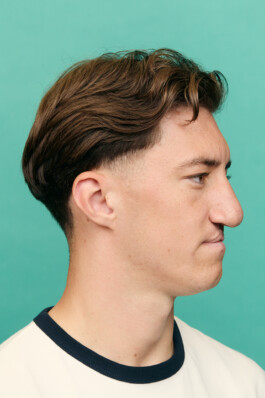 Angelo Stiller in profile, facing right, against a plain aqua background. He is wearing a cream-colored shirt with a dark navy collar. The image is closely cropped to highlight his facial features and hairstyle. Photo by Roderick Aichinger for Sports Illustrated Germany.