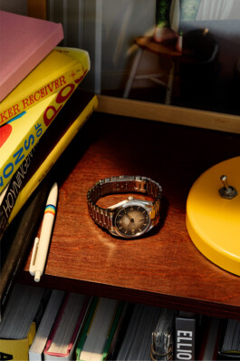 Photograph by Roderick Aichinger for Esquire Germany, fall 2025 issue. The image features an Omega Seamaster Railmaster 38mm (ref. 235.10.38.20.13.001) with a gradient brown dial and stainless steel bracelet, placed on a wooden surface between books and a yellow lamp. The scene highlights the updated, vintage-inspired design of the modern Railmaster in a creative, mid-century workspace setting.