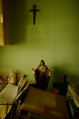 Small statues of Jesus and a saint, religious documents, and a crucifix on a green wall above a cluttered desk inside Schloss Goldenstein convent, Austria