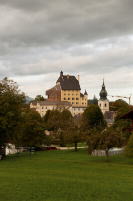 Exterior view of Schloss Goldenstein convent and church tower in Elsbethen, Austria, surrounded by trees and lawn under a cloudy sky.