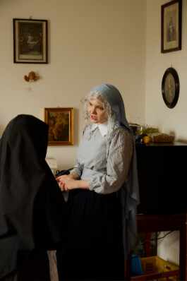 Younger woman with a blue headscarf holding hands and speaking with an elderly nun in a black habit, inside a room decorated with religious artwork.