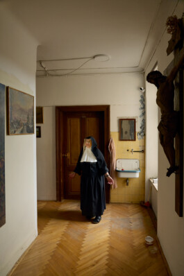 Sister Rita, an elderly nun in traditional habit, standing in a sunlit hallway near a wooden door and wall-mounted crucifix at Schloss Goldenstein convent in Austria.
