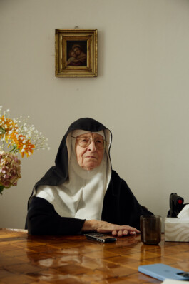 Sister Regina, an elderly nun in traditional black and white habit, seated at a wooden table with flowers and personal items, inside Schloss Goldenstein convent in Austria.