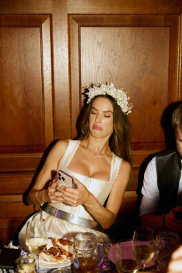 Brazilian model Alessandra Ambrosio, wearing a white dress and a flower crown, reacts to her phone while seated at a wooden table with food and drinks at Heidi Fest in Munich. Photo by Roderick Aichinger for The New York Times, 2025.