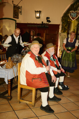 Wilfried Gliem and Wolfgang Schwalm of the Wildecker Herzbuben, dressed in matching Bavarian outfits and hats, entertain guests onstage with microphones at Heidi Fest in Munich. Other partygoers and servers in traditional clothing are visible in the background. Photo by Roderick Aichinger for The New York Times, 2025.