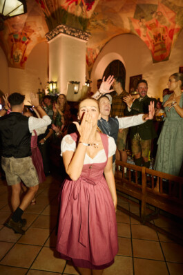 At the Hofbräuhaus in Munich during Heidi Fest, a woman in a pink dirndl stands in the foreground blowing a kiss towards the camera, while guests in traditional attire dance and celebrate in the festively decorated beer hall. Photo by Roderick Aichinger for The New York Times, 2025.