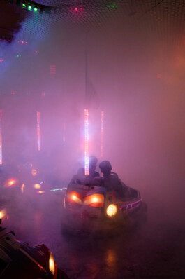 Two people ride together in a brightly lit bumper car, surrounded by fog and colorful lights at an Oktoberfest fairground in Munich. The hazy atmosphere creates a dreamy, immersive feel, with the silhouettes of other bumper cars and lights visible in the smoky background.