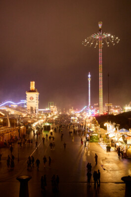A panoramic night view of Oktoberfest at Theresienwiese in Munich. The brightly lit fairground features crowds of people strolling along wet pathways, illuminated beer tents, food stalls, and colorful amusement rides, including a tall illuminated swing ride and a tower with a giant Paulaner beer stein on top. The festive atmosphere is captured under a moody, overcast sky.