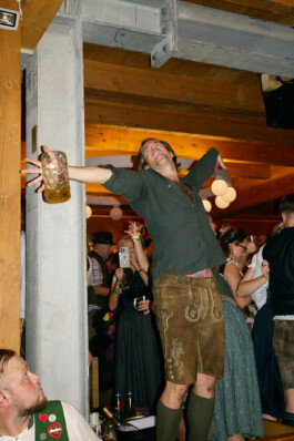 A lively scene at Oktoberfest in Munich shows a man standing on a bench or table, arms spread wide as he celebrates exuberantly. He wears traditional Bavarian lederhosen, dark socks, and holds a large beer stein. The background is filled with fellow festival-goers, some in dirndls and traditional attire, highlighting the joyful, raucous atmosphere of the beer tent.