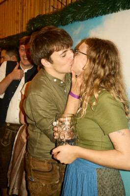 A man and woman in traditional Bavarian outfits share an affectionate moment at Oktoberfest 2025, with the woman holding a large beer stein and festivalgoers in traditional clothing in the lively beer hall atmosphere.