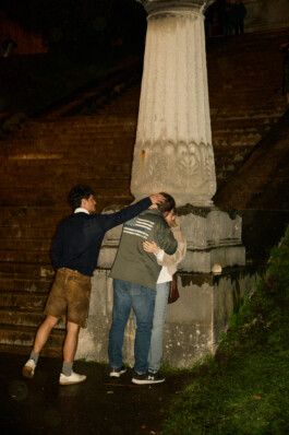 At night, at the foot of the stairs leading to Munich's Bavaria statue at Theresienwiese, a person in lederhosen drunkenly pushes a man's head towards a woman, both standing next to a stone column. The man wears a jacket with an altered American flag, referencing right-wing symbolism and protest. The candid, chaotic moment reflects both Oktoberfest's festive and sometimes political atmosphere.