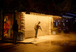 Late at night at Oktoberfest in Munich, a man uses a pressure washer to clean the front of a brightly lit, closed fairground booth. Steam rises around him, illuminating the scene, while another man stands nearby watching. The foreground shows wet pavement, suggesting the end of a lively festival night and the start of cleanup.