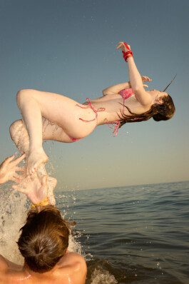 Teenage girl in a pink bikini is flipped into the air above the water by a friend during a lively swim at the beach, with sunlit splashes and sky in the background. Dynamic, joyful summer scene by Roderick Aichinger for Spiegel Magazine's youth camp article.