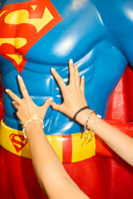 Close-up of a teenage girl's hands pressing against the chest of a large Superman statue, highlighting playful interaction and colorful bracelets. Captured by Roderick Aichinger during a summer camp day trip to Siena for Spiegel Magazine's youth camp story.