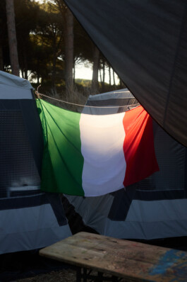 Italian flag hangs between tents, illuminated by sunlight during golden hour at a pine-shaded summer youth camp near Tarquinia. Symbolic camp scene shot by Roderick Aichinger for Spiegel Magazine