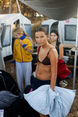 Teenage girl in a brown bikini top and blue skirt stands inside a youth camp tent, holding a striped pillow and wearing a red backpack. Other campers and tents are visible in the background, capturing the end of a week-long summer youth camp near Tarquinia. Photo by Roderick Aichinger for Spiegel Magazine.