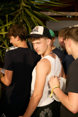 Flash-lit photo of teenage boys standing in line for a meal at a summer youth camp, one wearing a white tank top and trucker hat, holding a plate. The group waits together under late afternoon light near tropical plants. Photo by Roderick Aichinger for Spiegel Magazine, near Tarquinia.