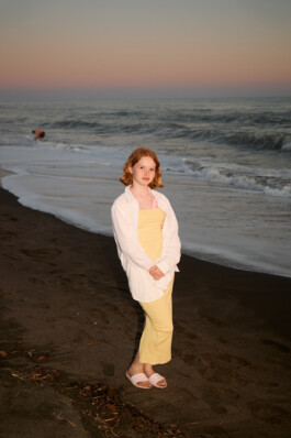 Teenage girl with short red hair stands on a dark beach at sunset, wearing a yellow dress, open white shirt, and pink slide sandals. The subject is brightly illuminated with direct flash, making her stand out against the pastel sky and waves. Documentary photo by Roderick Aichinger from a summer youth camp near Tarquinia for Spiegel Magazine.