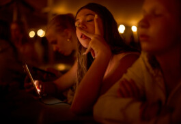 Warmly lit, candid night photo of a teenage girl at summer camp leaning on a table, intently recording or editing TikTok content on her phone. The screen glows in the dim, orange light with other teenagers sitting nearby. Photo by Roderick Aichinger for Spiegel Magazine, youth camp near Tarquinia.