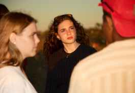 Flash-lit sunset photo of teenage friends talking in a group on the beach at summer camp. The central girl with curly hair and sunglasses is warmly illuminated, with other teens partially visible at the edges. Photo by Roderick Aichinger for Spiegel Magazine, youth camp near Tarquinia.