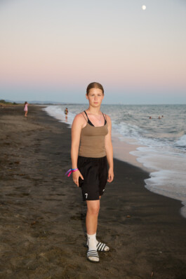 Flash-lit daytime photo of a teenage girl at summer camp, standing outside tents, holding a small mirror up to her face while using an eyelash tool to do her lashes. She wears a blue tank top and bracelets. Photo by Roderick Aichinger for Spiegel Magazine, youth camp near Tarquinia.
