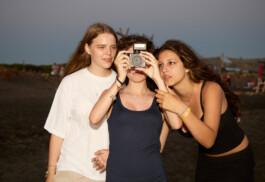 Flash-lit sunset photo of three teenage girls on the beach at summer camp, one holding a compact camera with a flash while her friends gather close and look on. Casual scene of friendship and summer evening. Photo by Roderick Aichinger for Spiegel Magazine, youth camp near Tarquinia