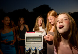 Flash-lit photo at dusk showing a group of teenage girls at a summer camp. Several girls gather around a large outdoor power outlet box, using it as a stand for their mobile phones while shooting TikTok content. Trees are silhouetted in the background. Photo by Roderick Aichinger for Spiegel Magazine, youth camp near Tarquinia.