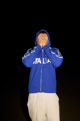 Flash-lit night portrait on the beach of a teenage boy wearing a blue 'Italia' hoodie and light sweatpants, standing against a pitch-black background with his hands covering his face. Candid documentary photo by Roderick Aichinger for Spiegel Magazine, summer youth camp near Tarquinia.