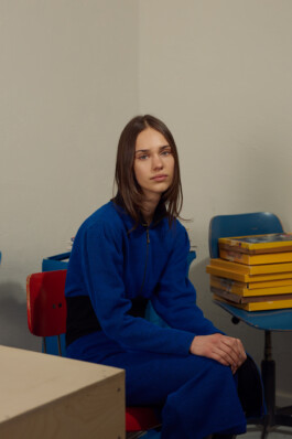 Young woman with shoulder-length brown hair sits thoughtfully on a red chair during a lighting test, wearing a royal blue zip-up jumpsuit with black waistband. She leans forward with hands on knees beside a small blue table stacked with yellow boxes, set against a plain wall with a wooden stool nearby.