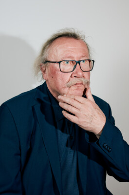 Peter Sloterdijk poses in a dark blue suit against a plain light background, his hand raised thoughtfully to his chin in a formal portrait at Schloss Elmau.