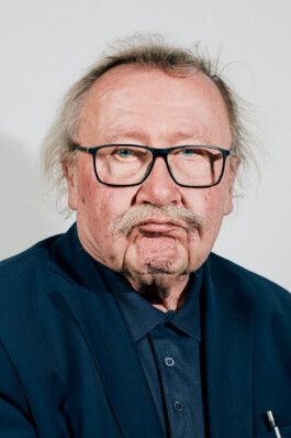Peter Sloterdijk poses in a dark blue suit against a plain light background, his hand raised thoughtfully to his chin in a formal portrait at Schloss Elmau.