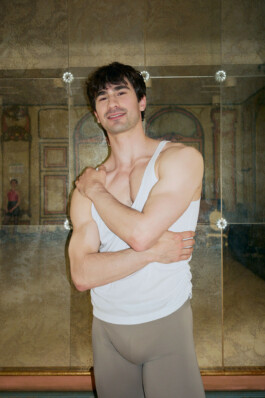 Portrait of Clark Eselgroth, dancer with Bayerisches Staatsballett, standing in a white tank top in front of a mirrored wall.