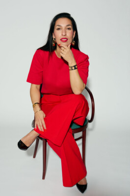Tijen Onaran sits on a chair in a red outfit against a white background.
