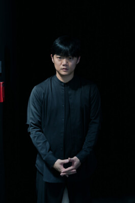 Portrait of choreographer Lai Hung Chung standing against a black background, wearing a dark button-up shirt with his hands clasped in front of him.