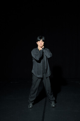 Choreographer Lai Hung Chung standing on a dark stage with his legs apart and hands around his neck, wearing a black shirt, loose trousers, and sneakers.