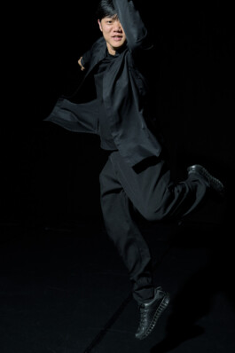 Choreographer Lai Hung Chung leaping across a dark stage, one arm raised and one leg bent behind him, wearing a black shirt, loose trousers, and sneakers.