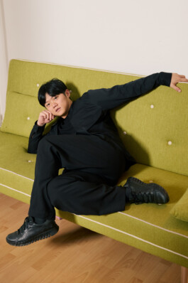 Choreographer Lai Hung Chung reclining diagonally on a lime-green sofa, wearing black clothing and black sneakers, with one arm extended along the backrest.