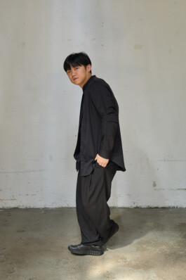 horeographer Lai Hung Chung standing in profile against a worn light-gray wall, wearing a loose black shirt, wide-leg trousers, and black sneakers.